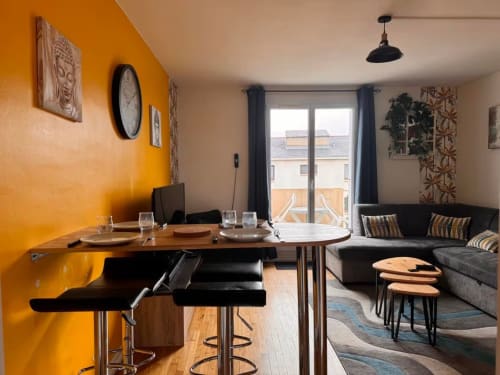 Studio moderne à Val'Déco Conciergerie avec mur d'accent orange, coin repas, kitchenette et canapé gris avec vue sur Les Ponts