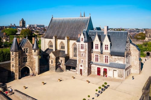 Cour de château médiéval à Les Ponts de Cé avec murs de pierre, portes rouges et toits d'ardoise sous ciel bleu