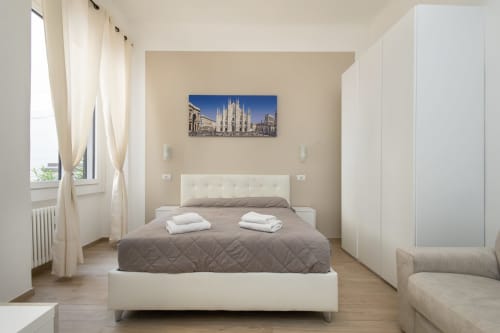 Modernes Schlafzimmer in den Magnolia Apartments Mailand mit cremefarbenem Polsterbett, grauer gesteppter Bettdecke, Kunstwerk der Mailänder Kathedrale