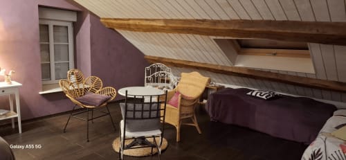 Chambre cosy sous les toits de La Maison de Marie-Claire avec murs violets, fauteuil en osier et vue sur le jardin