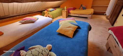 Chambre mansardée colorée à La Maison de Marie-Claire avec lit bleu, coussins colorés, plafond en bois et jouets.