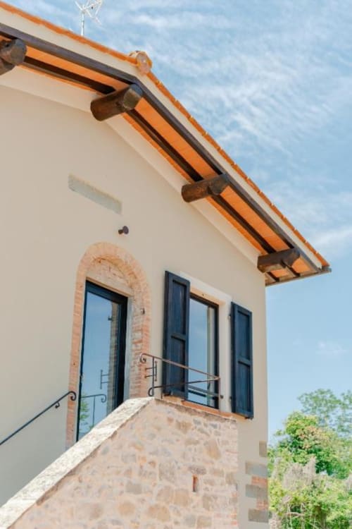 Esterno rustico toscano con portale ad arco, persiane scure, travi in cotto, balconata e vista sul giardino