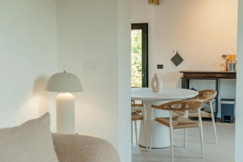 Luminosa sala da pranzo nell'appartamento Barn di Tenuta Alela con tavolo rotondo bianco, sedie in vimini e vista sul giardino dalla porta.