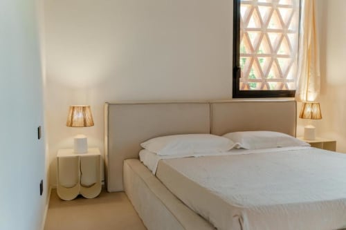 Camera minimalista a Tenuta Alela con letto in tessuto beige, biancheria bianca, lampade a sospensione intrecciate e vista giardino