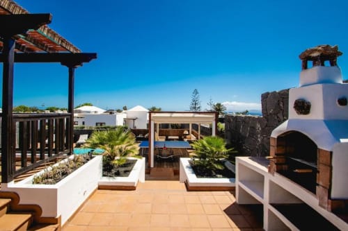 Terraza en la azotea de Villa Cavendish en Playa Blanca con edificios encalados, pérgola de madera oscura, plantas de palmera y vistas al océano.