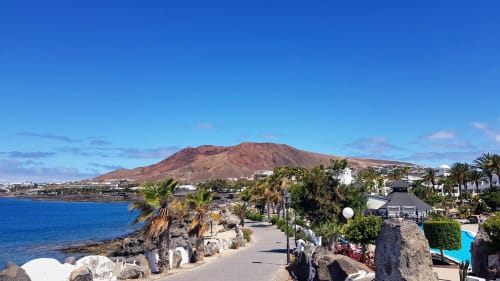 Paseo frente al mar de Villa Cavendish con palmeras, montaña volcánica y océano azul en Playa Blanca