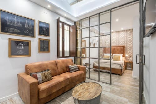 Estudio CTH Granada Loft Gracia con sofá de cuero coñac, división de vidrio, cama y arte mural de planos