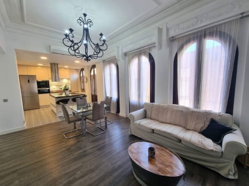 Sala de estar de la Penthouse Gracia en CTH Granada con mobiliario moderno, cocina, mesa de comedor, sofá beige y ventanales arqueados