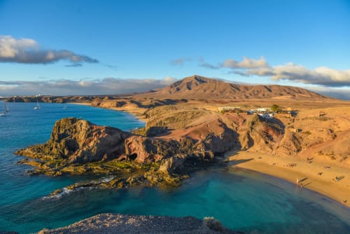 Aguas turquesas y acantilados dorados en Playa Blanca, Lanzarote, con montaña volcánica de fondo y veleros