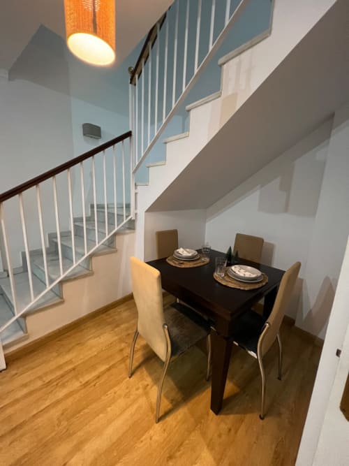 Comedor moderno con mesa negra, sillas color crema, suelos de madera y escalera en apartamento de Jerez de la Frontera