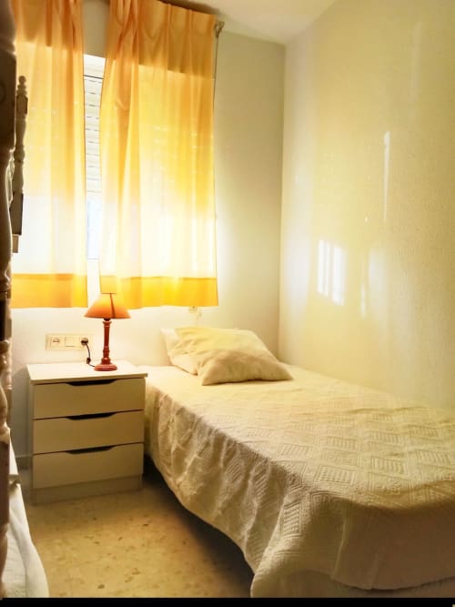 Dormitorio luminoso en Top Home Integra Jerez con cama individual, mesilla blanca, lámpara naranja y cortinas amarillas