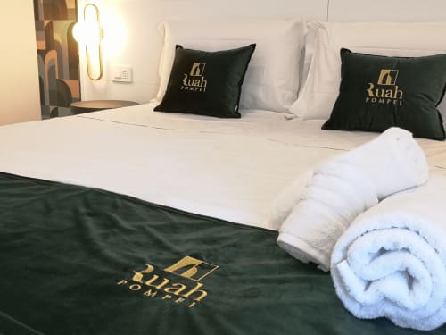 Ruah Pompei hotel room with white bed, black pillows, rolled towels, and dark blanket with gold logo
