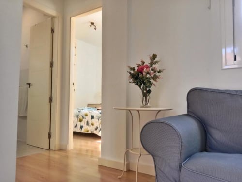 Dormitorio moderno con sillón azul, mesita con flores rosas y puerta del baño abierta en Jerez de la Frontera