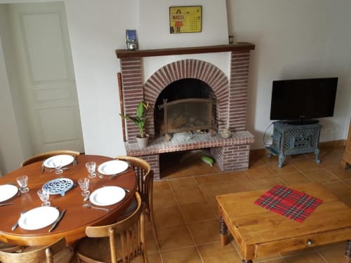 Dining room with fireplace at Le Petit Logis, Avranches. Round wooden table, brick hearth, TV, terracotta floor.