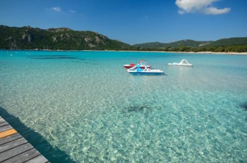 Baia turchese cristallina presso le Villas Lantana a Lecci con barche ormeggiate, spiaggia sabbiosa e colline boscose.