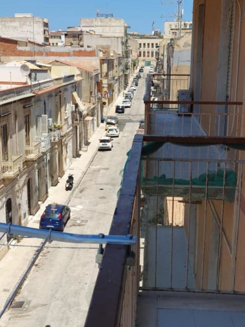 Vista dal balcone di una strada di Siracusa con auto parcheggiate e storici edifici gialli sotto un cielo azzurro limpido