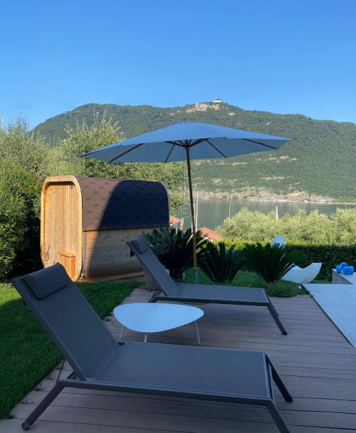 Verdolivo lakeside retreat with lounge chairs, blue umbrella, and wooden barrel sauna overlooking mountain views
