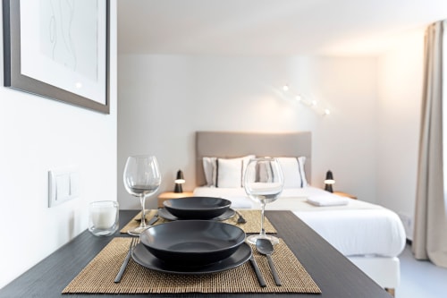 Elegante estudio en hotel de Oporto con zona de comedor, platos negros, copas de vino y cama con cabecero neutro