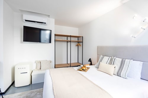 Modern studio bedroom in Porto hotel with wall-mounted TV, wooden closet, gray upholstered bed, and compact kitchenette.