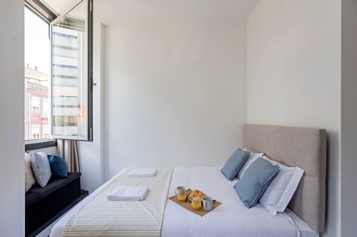 Compacta habitación estudio en hotel de Porto con vistas a la ciudad, cama queen con almohadas azules, asiento junto a la ventana y bandeja de desayuno