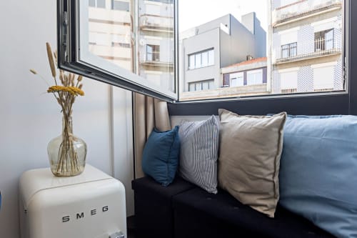 Ventana de estudio compacto con vistas a la ciudad, cama con almohadas, mesita blanca con flores secas en jarrón de vidrio, Oporto