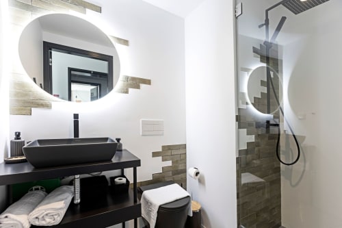 Baño moderno en el estudio Benefit Collection Porto con lavabo negro, espejo circular iluminado y ducha abierta