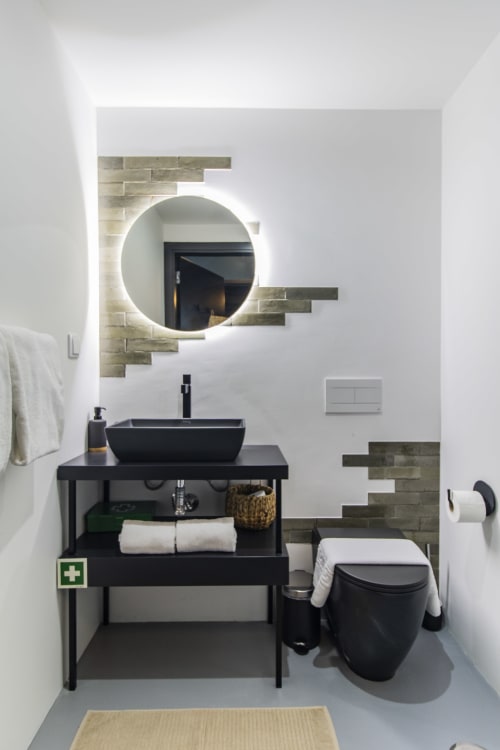 Baño moderno en hotel de Porto con lavabo negro suspendido, espejo circular iluminado, acentos de piedra y inodoro negro