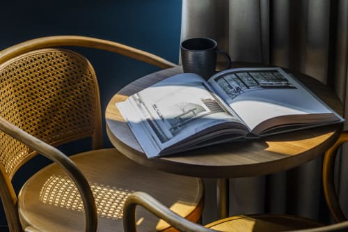 Espacio de trabajo iluminado por el sol en habitación de hotel oporto con revista de arquitectura abierta, silla de rattán, taza oscura y vista al jardín