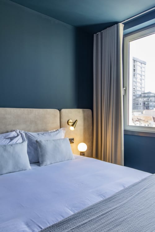 Habitación de hotel moderna con paredes verde azulado, cabecero beige, ropa de cama azul y gran ventana con vistas a Oporto en Benefit Collection