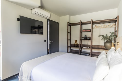 Moderno y compacto dormitorio estudio en hotel de Porto con cama blanca, TV montada en pared, estanterías de madera, aire acondicionado y