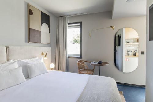 Modern compact studio bedroom in Porto hotel with white bed, wooden desk, round mirror, and kitchenette visible in background