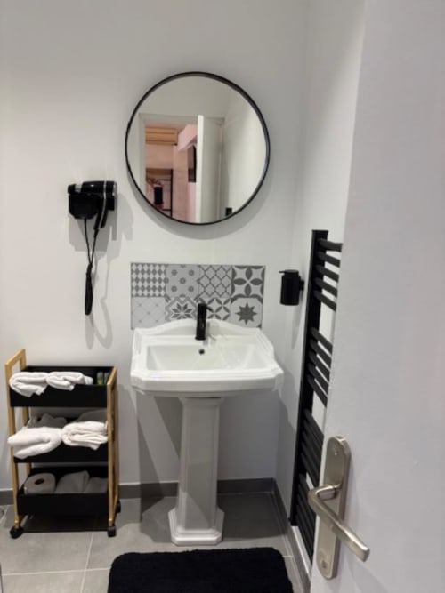 Salle de bains moderne de l'Hôtel Manon à Marseille avec lavabo sur piédestal, miroir rond, sèche-serviettes chauffant et rangements.