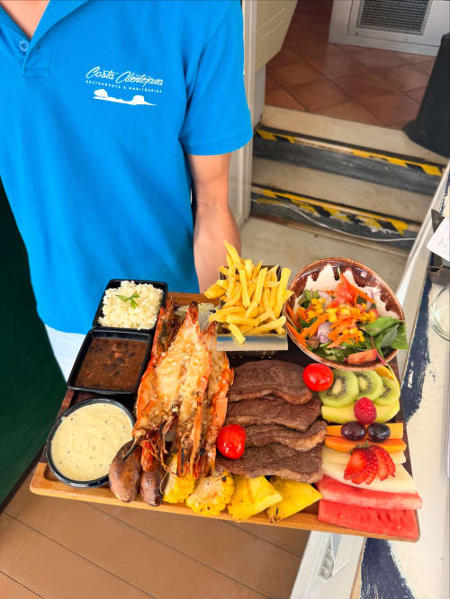 Staff member at Costa Alentejana holding wooden platter with grilled shrimp, steak, fries, salads and fresh fruit