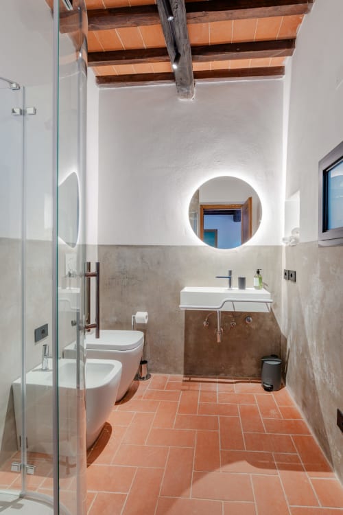 Bagno moderno a Tenuta Alela con piastrelle in cotto, specchio circolare illuminato, lavandino bianco, bidet, wc e dettagli in legno