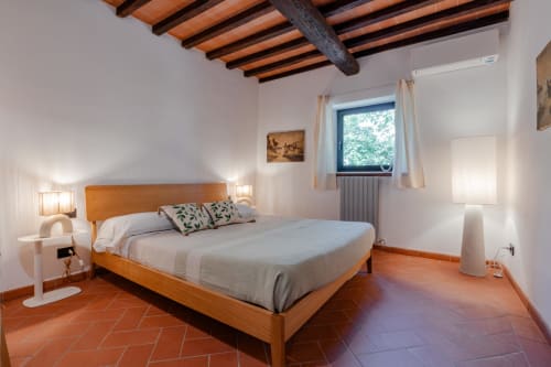 Camera luminosa a Tenuta Alela con letto in legno, pavimento in cotto, travi a vista e finestra con vista sul giardino.
