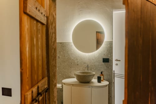 Bagno moderno con specchio rotondo illuminato, lavabo in pietra e mobile in legno presso l'appartamento di lusso Tenuta Alela