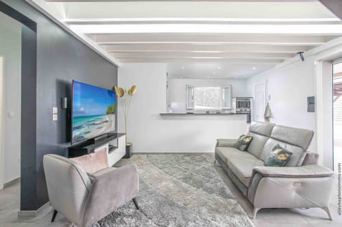 Modern villa living room with gray sofa, accent chair, 65-inch TV, beach scene art, kitchen area, and contemporary decor in