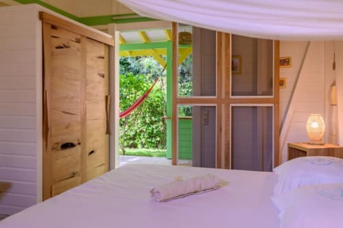 Chambre du gîte Ti'Kaz Lime avec vue jardin, portes en bois, volets verts, lampe tressée et accès à la terrasse