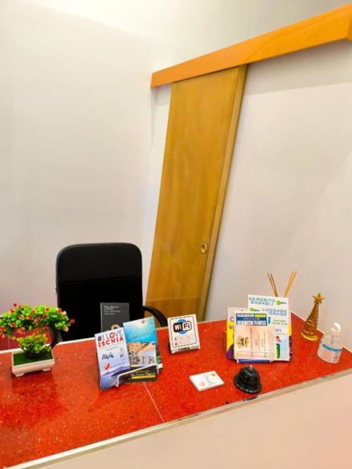 Bancone alberghiero con piano rosso, asta tenda gialla, fiori in vaso, cartello WiFi e brochure turistiche presso I Migliori Artisti in