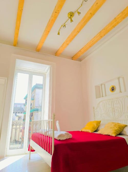 Luminosa camera da letto a I Migliori Artisti, Napoli, con biancheria rossa, cuscini gialli, travi in legno e vista sulla città dal balcone