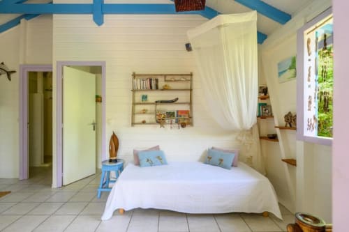 Chambre lumineuse au cottage Ti'Kaz Cannelle à Marie-Galante avec lit blanc, poutres bleues, voile de lit et vue sur le jardin