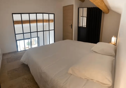 Bright bedroom with white bedding and wooden beam in Provençal cottage, featuring gridded window and black door.