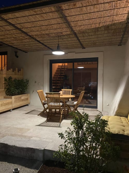 Night view of Mas St André cottage patio with wooden dining table, chairs, reed ceiling, and glass doors opening to interior,