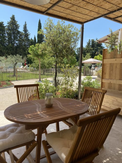 Wooden dining table with chairs under pergola at Mas St André cottage in Provence, overlooking gardens and pool area