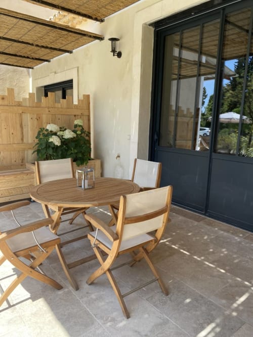 Outdoor patio with wooden round table and chairs, white cushions, flowering planter, black glass doors, and woven pergola at