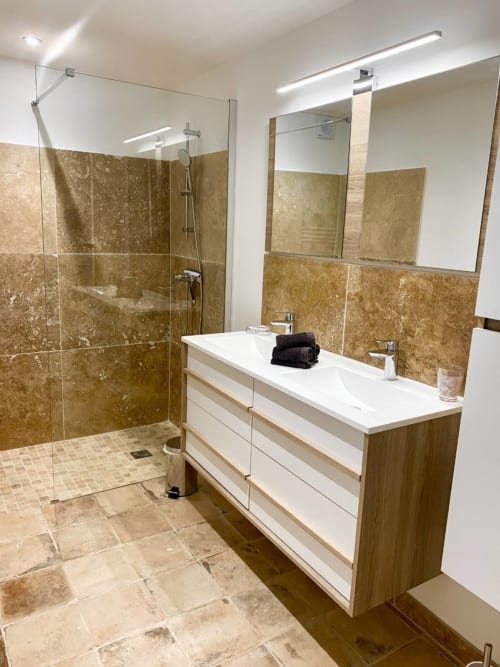 Modern bathroom in Provence cottage with travertine tile, glass shower, white vanity, and mirrors with lighting at Mas St