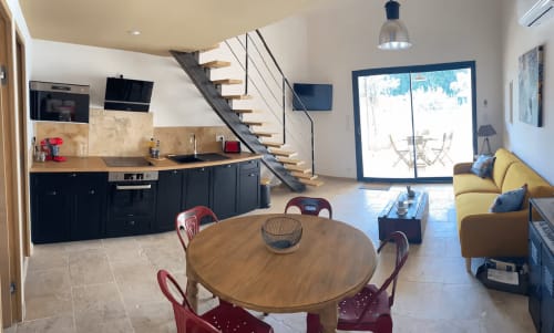 Modern open-plan cottage kitchen and dining area with wooden table, black cabinetry, and wooden staircase at Gîtes Olivier in