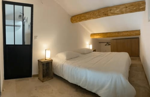 Modern bedroom in Provence cottage with exposed wooden beams, white bedding, black-framed glass door, and warm ambient