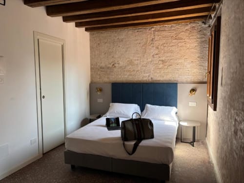 Camera moderna di Casanova Hideaway Venice con testata scura, biancheria bianca, mattoni a vista e travi in legno