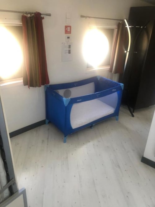 Blue travel crib with white mattress in bright Family Studio at Cosyade hotel, Bourbourg, with windows and red curtains
